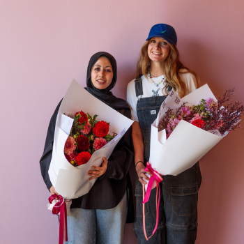 Two women standing against a pink wall, each holding a wrapped bouquet of red and pink flowers tied with ribbon.